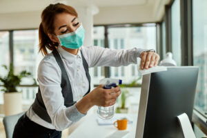 Young businesswoman with face mask disinfecting her desktop PC while working in the office during coronavirus epidemic.